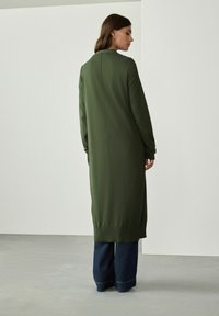 Lang grøn cardigan i et blødt stof med lige snit, der har ribbede manchetter og en søm ned ad ryggen, båret over blå brede jeans.