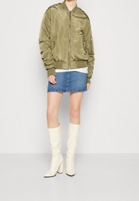 Olivgrön satinbombarjacka med ribbad muddar, i kombination med en denimminikjol. Modellen bär vita knähöga blockklackstövlar.