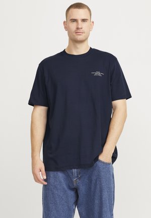 Camiseta de algodón azul marino con un corte relajado, mangas cortas, cuello redondeado y un pequeño detalle de texto blanco en el pecho. Llevada con jeans azules.