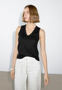 Top senza maniche di colore nero con scollatura a V, realizzato in un tessuto liscio. Presenta un bordo a contrasto ed è abbinato a pantaloni bianchi a vita alta.