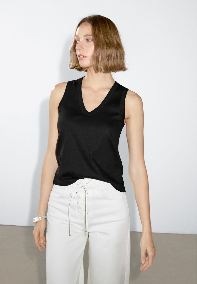 Top senza maniche di colore nero con scollatura a V, realizzato in un tessuto liscio. Presenta un bordo a contrasto ed è abbinato a pantaloni bianchi a vita alta.