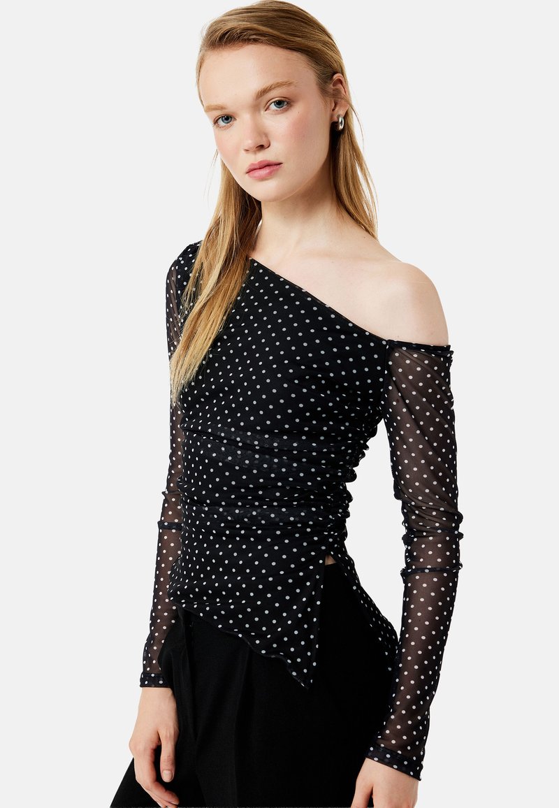 Sort off-shoulder top med hvide polkaprikker, med rynkede detaljer og gennemsigtige lange ærmer, afsluttet med en asymmetrisk nederkant.