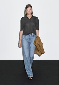 Czarna, prążkowana koszula polo, jasnoniebieskie dżinsy z wysokim stanem, trzymająca jasnobrązową, zamszową kurtkę. Czarne sandały o minimalistycznym designie.