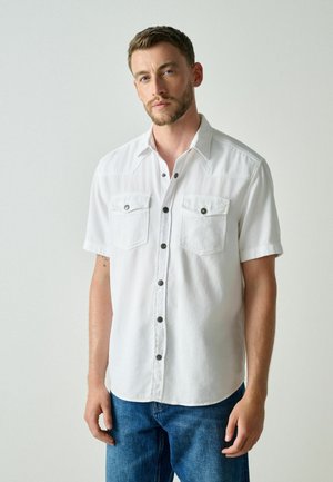 Man with short hair and beard wearing a white short-sleeve button-up shirt and blue jeans, standing against a plain light background.