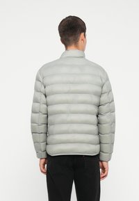 Polstret grå jakke med en myk tekstur, med stående krage og horisontal quiltedesign, sett fra baksiden.