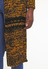 Stickad lång kappa i svart och senapsgul med strukturerade mönster och lös passform. Har ribbade kanter och en öppning fram.