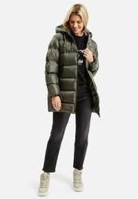 Oliwkowa kurtka puchowa z kapturem, z zamkiem błyskawicznym z przodu, pikowany wzór i luźny krój. W zestawie z czarną koszulą i ciemnymi dżinsami.