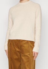 Ljusbeige ulltröja med rund halsringning, ribbade ärmslut och nederkant; parat med bruna manchesterbyxor med vertikala ränder.