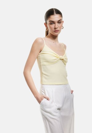 Keltainen teksturoitu crop-top, jossa on kierretty etuosa, ohuet hihnat, yhdistettynä korkeavyötäröisiin valkoisiin housuihin. Malli seisoo kädet taskuissa.