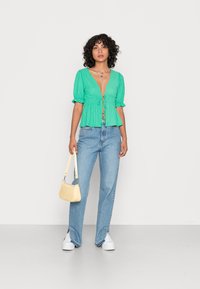 Blusa verde texturizada com mangas bufantes, decote em V profundo e detalhe de laço. Combinada com jeans de perna larga azul claro e uma pequena bolsa amarela.
