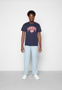 T-shirt de algodão azul marinho com o logótipo "TOMMY JEANS" em vermelho, combinado com calças de ganga azul claro e ténis brancos, contra um fundo neutro.