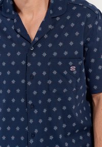 Chemise à manches courtes de couleur marine avec des motifs floraux blancs et turquoise, dotée d'une poche poitrine et d'un col à boutonnière. Fabriquée en tissu léger.