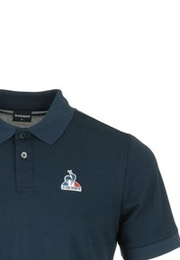Polo de algodón azul marino con una tapeta de tres botones y logotipo bordado que presenta un gallo en colores rojo, blanco y azul.