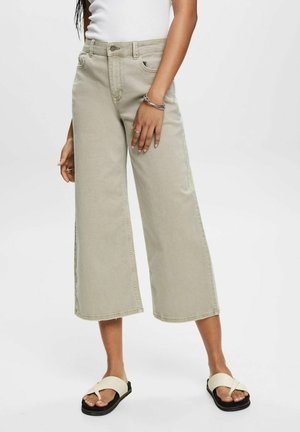 Lichtbeige wijde jeans met hoge taille, voorzien van voorzakken en een lange zoom. Gecombineerd met lichtgekleurde platte sandalen.