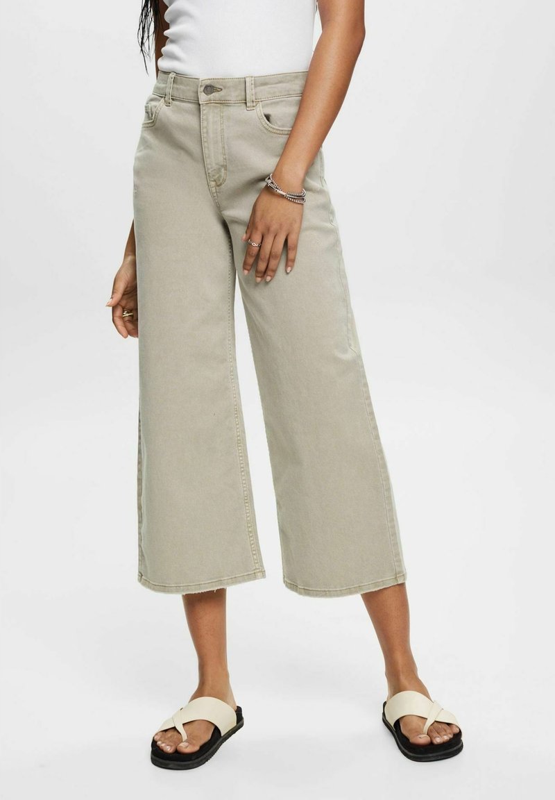 Jean large couleur beige clair avec taille haute, doté de poches avant et d'un ourlet long. Porté avec des sandales plates de couleur claire.