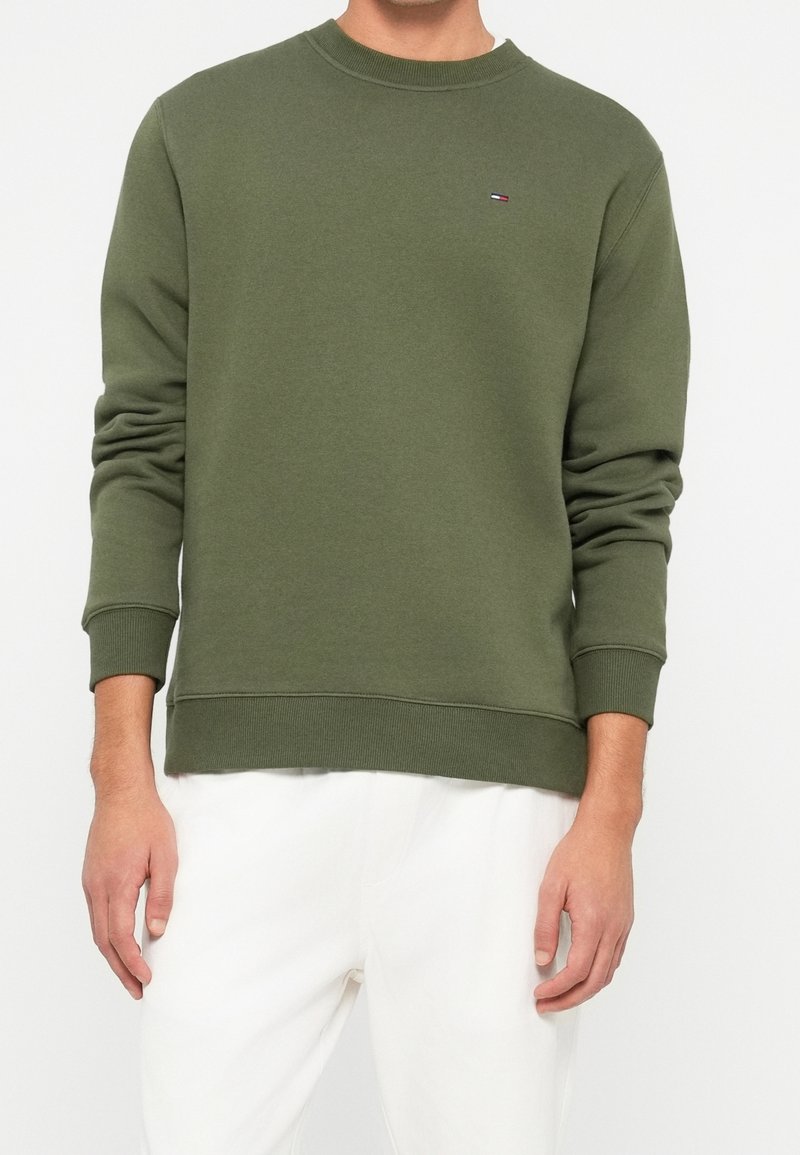 Homme portant un sweat-shirt vert olive à col rond avec un petit logo sur la poitrine et un pantalon blanc, debout devant un fond uni.