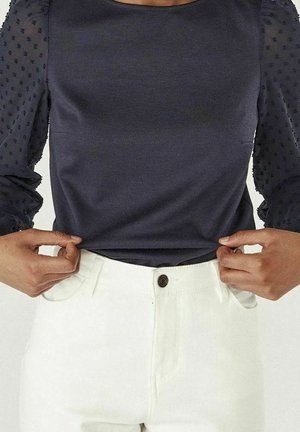 Osoba w granatowej bluzce z przezroczystymi, w kropki rękawami i białych, wysokich spodniach, trzymająca dolną część bluzki obiema rękami.