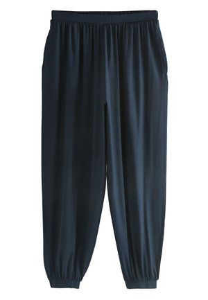 Persona con pantalones oscuros y sueltos con cintura elástica, mano dentro del bolsillo derecho, camisa azul metida dentro.