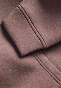 Primo piano del tessuto di un maglione marrone, con un polsino piegato, texture liscia e dettagli di cucitura visibili. Design complessivo semplice e minimale.