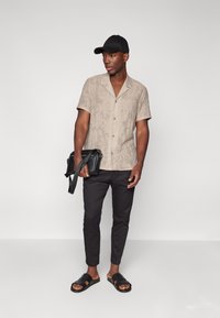 Camisa beige de manga corta con un patrón de hojas, pantalones negros, gorra negra y sandalias negras. Sosteniendo un bolso cruzado negro.