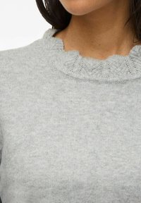 Närbild av en person som bär en ljusgrå stickad tröja med en vågig, ribbad halsringning och långt mörkt hår synligt.