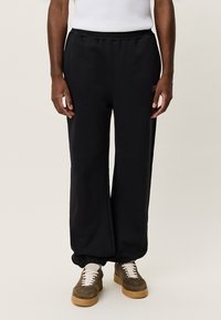 Μαύρο παντελόνι φόρμας από μαλακό ύφασμα, με ίσια κοπή, ελαστική μέση και μανσέτες στους αστραγάλους, σε συνδυασμό με καφέ αθλητικά παπούτσια.