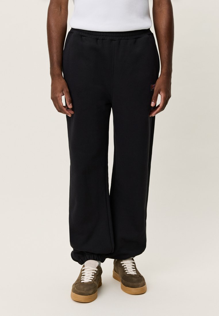 Μαύρο παντελόνι φόρμας από μαλακό ύφασμα, με ίσια κοπή, ελαστική μέση και μανσέτες στους αστραγάλους, σε συνδυασμό με καφέ αθλητικά παπούτσια.