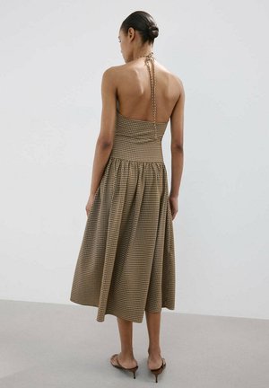 Mujer con el pelo recogido en un moño, vistiendo un vestido midi marrón de cuello halter con un pequeño patrón de cuadros, combinado con tacones marrones de espalda abierta, de perfil.