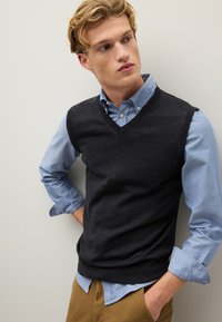 Dunkelgraue Wollweste mit V-Ausschnitt, über ein hellblaues Hemd mit Knöpfen getragen. Der Stoff wirkt glatt; subtile Texturdetails sind sichtbar.