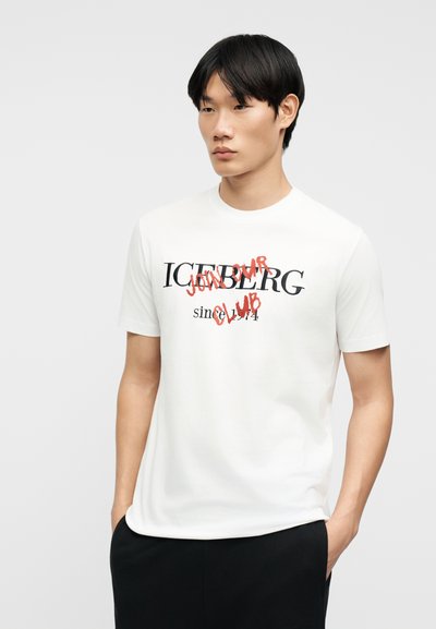 Νεαρός άνδρας που φοράει λευκό μπλουζάκι με μαύρο και κόκκινο κείμενο που αναγράφει "ICEBERG since 1974 JOIN OUR CLUB", με τα χέρια στις τσέπες.