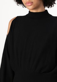 Black knitted sweater featuring a high neck, open shoulder design, and gathered waist. Soft texture with ribbed accents near the collar and hem.