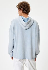 Sudadera azul de gran tamaño con un acabado suave y texturizado. Presenta capucha con cordón y mangas amplias, combinada con pantalones blancos.