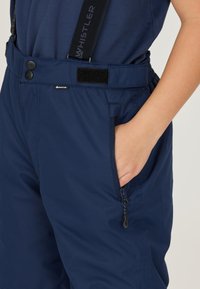 Mörkblå snöbyxor med justerbara band, två framfickor, en ficka med dragkedja och en mjuk tygtextur. Har en svart velcro-accent.