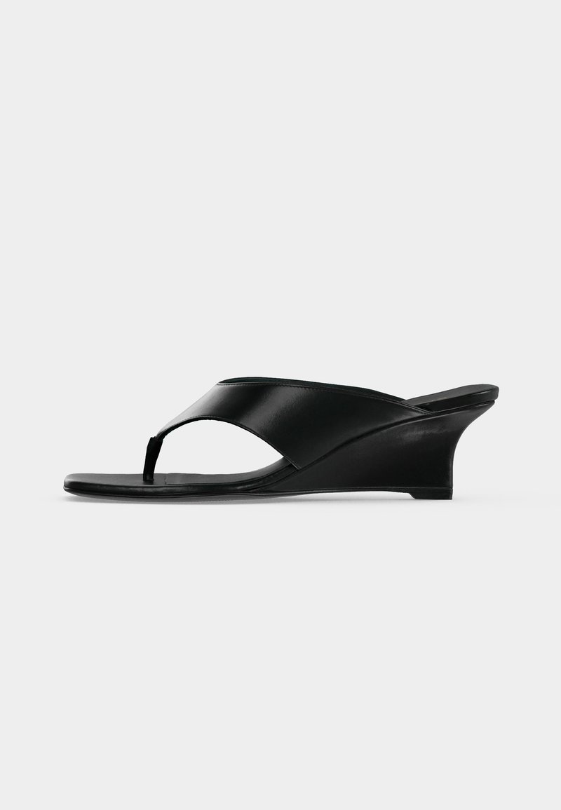 Sandale compensée en cuir noir avec bride entre les orteils et bout ouvert, dotée d'un talon bas incurvé et d'un design minimaliste épuré.