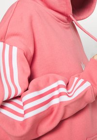 Růžová mikina s měkkou texturou, s bílými pruhy na rukávech a kapucí na šňůrku. Obsahuje logo na přední straně.