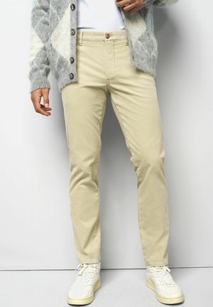 Beige Baumwollhosen mit schmaler Passform. Mit Knopfverschluss und Fünf-Taschen-Design. Getragen mit einer strukturierten grauen Strickjacke und weißen Turnschuhen.