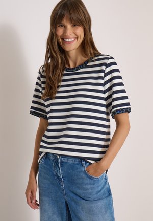 Kobieta uśmiecha się, ubrana w czarno-biały pasiasty t-shirt z krótkim rękawem ozdobiony niebieskim wzorem i niebieskie dżinsy, z jedną ręką w kieszeni.