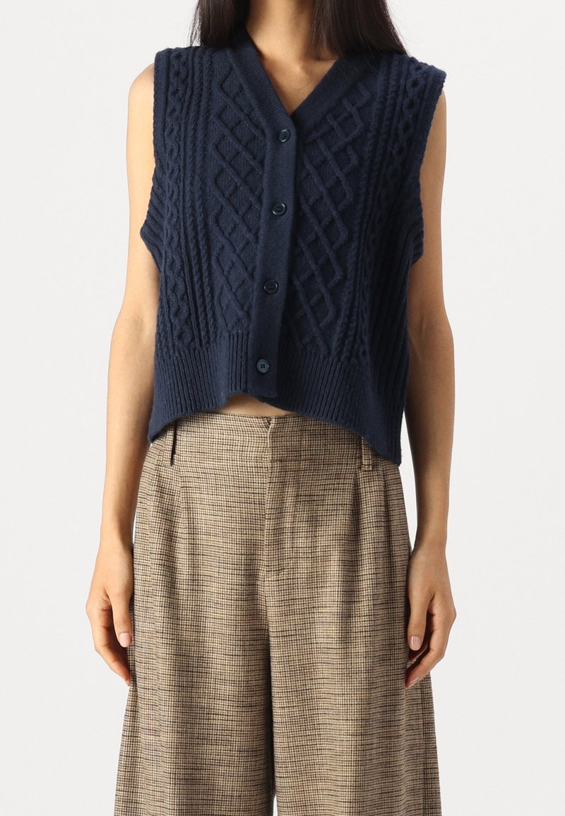 Veste sans manches bleu marine en maille torsadée avec boutons, portée sur un pantalon large à taille haute à carreaux marron.