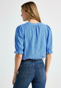 Kvinna i en blå blus med hålmönster och puffärmar samt mörkblå högmidjade jeans, sedd bakifrån mot en enfärgad bakgrund.