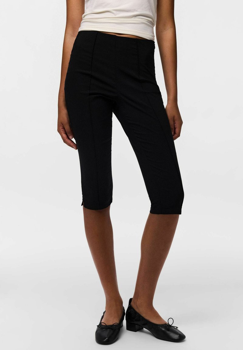 Femme portant un pantalon noir ajusté au genou, des chaussures noires à talons bas, et un haut blanc sans manches, debout devant un fond uni.