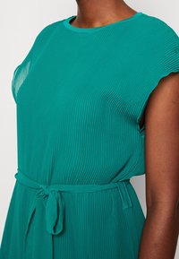 Teal, plisserad klänning med korta ärmar och rund halsringning. Har ett knutet bälte i midjan för extra definition. Mjuk, strukturerad tyg.