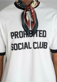Biela tričko s čiernymi lemovanými rukávmi s odvážnym čiernym textom "PROHIBITÉ KLUB SOCIÁLNEHO ZÁKAZU." Štýlový hodvábny šál uzle na krku.