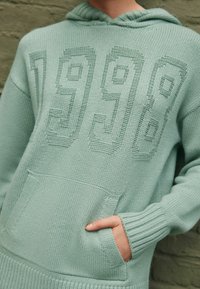 Miętowo-zielona dziana bluza z kapturem, z przednią kieszenią, ściągaczami na rękawach oraz teksturowanym wzorem "1998" na piersi. Miękka, średniej wagi tkanina.