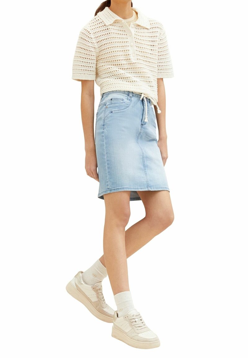 Femme portant un haut blanc en crochet à manches courtes, une jupe en jean bleu clair avec cordon, des chaussettes blanches et des baskets beiges, debout avec une jambe croisée.