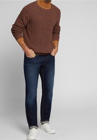 Brauner Zopfstrickpullover mit Rundhalsausschnitt, kombiniert mit dunkelblauen Slim-Fit-Jeans und weißen Sneakers. Rippstrickdetails an den Bündchen und dem Saum.