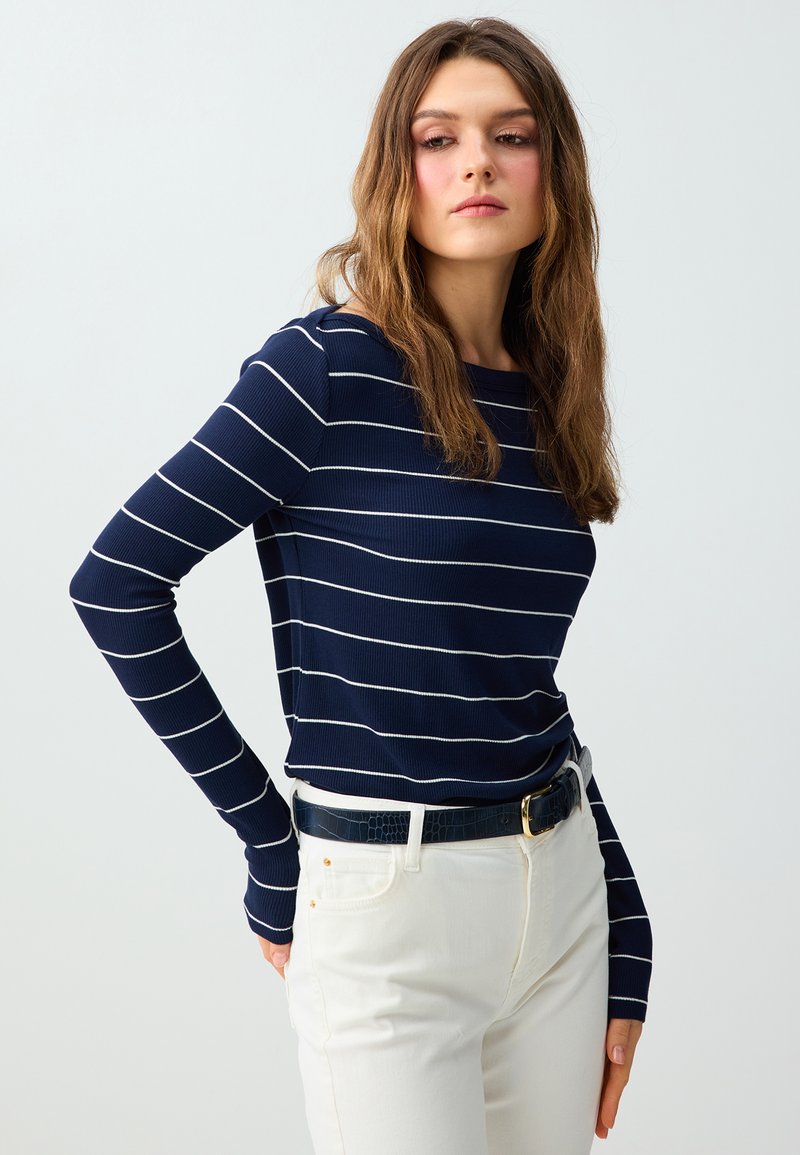 Femme aux longs cheveux bruns portant une chemise bleu marine à manches longues avec des rayures blanches horizontales et un pantalon blanc avec une ceinture noire, posant de profil.