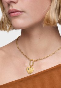 Collar de cadena de oro con un colgante en forma de corazón adornado con piedras preciosas en azul y rosa, sobre un fondo marrón texturizado.