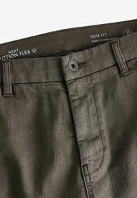 Pantalones ajustados en una tela de verde oliva oscuro, con cierre de botón, pasadores para cinturón y una textura sutil. La etiqueta indica diseño "Motion Flex".
