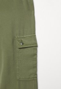 Sezione di pantaloni cargo verde oliva con tasca laterale con bottone, tessuto liscio e cuciture rinforzate.