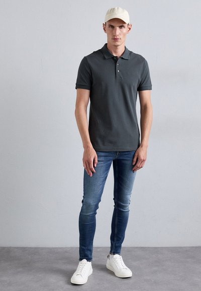 Pelēks polo krekls ar apkakli, tumši zili pieguļoši džinsi un baltas sporta kedas. Modelis valkā gaismas bēšas cepuri. Vienkārša, ikdienišķa apģērba kombinācija.